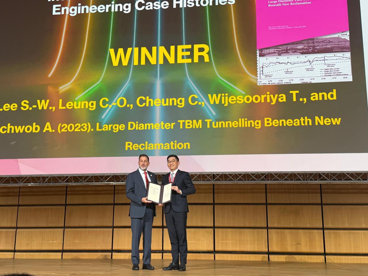 Two men standing on a stage holding an award Description automatically generated